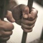 Man's hands tightly holding the bars of his jail cell.